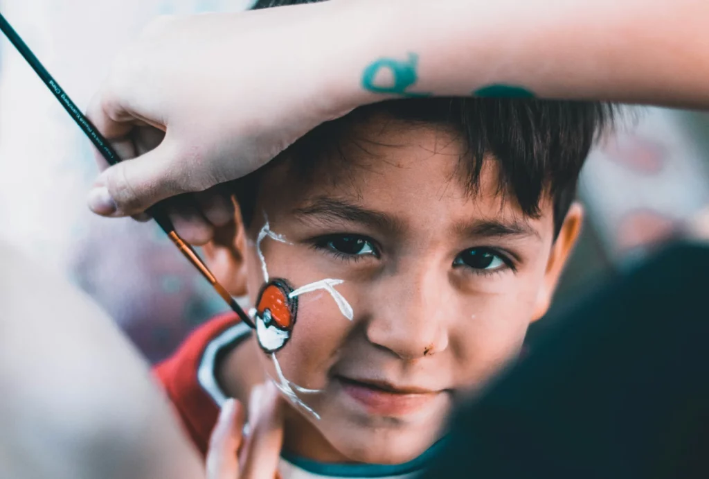 A face painter completing a Poké ball design on a child's face in face paint.