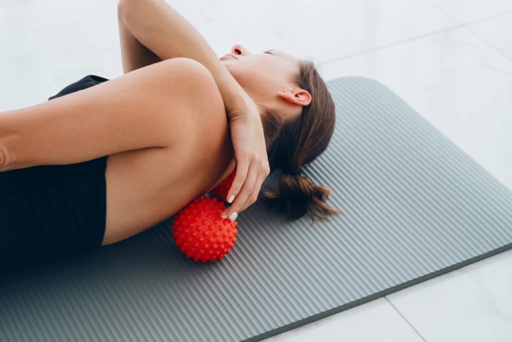A woman laying on a mat, demonstrating how to use a spikey massage ball to relieve your own tight shoulder and back muscles.