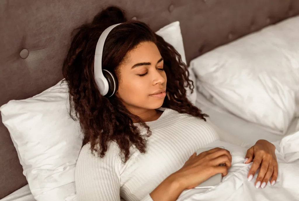 A woman wearing a white long-sleeve top and silver headphones meditates and holds her phone while reclining in a bed with a gray headboard and white bedding.