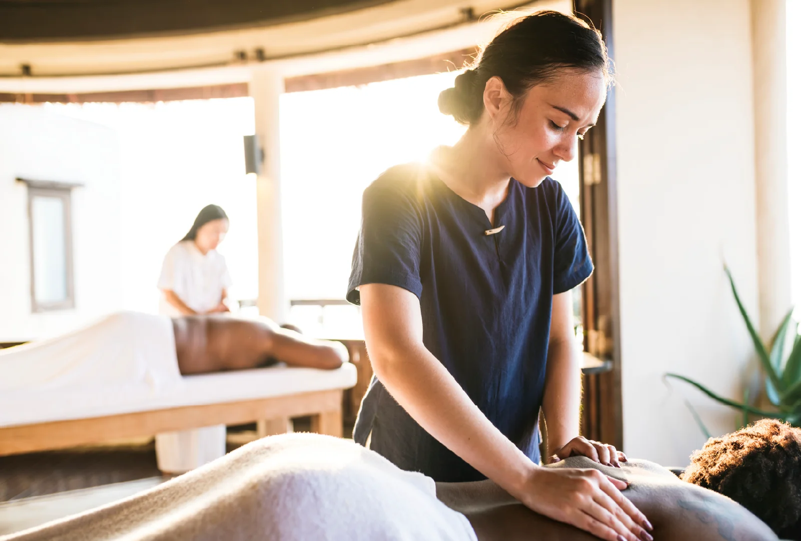A female massage therapist wearing a blue uniform performs a back massage on a client in a spa as sunlight streams in behind her.
