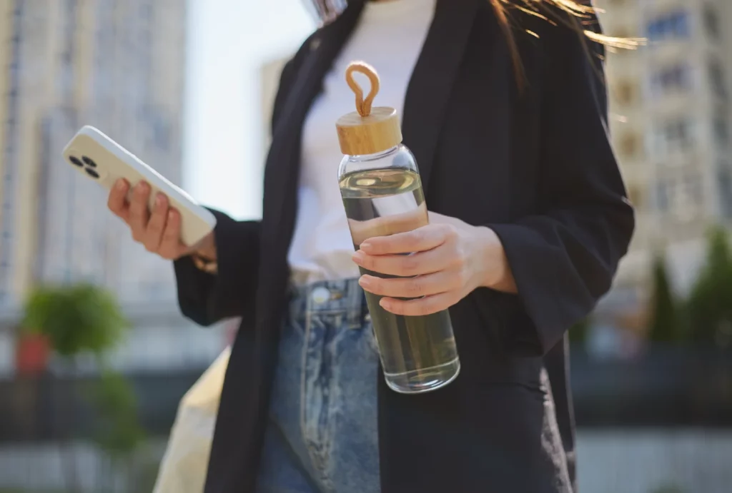 A person in business attire holding a phone in one hand and a reusable water bottle in the other, outdoors in the city.
