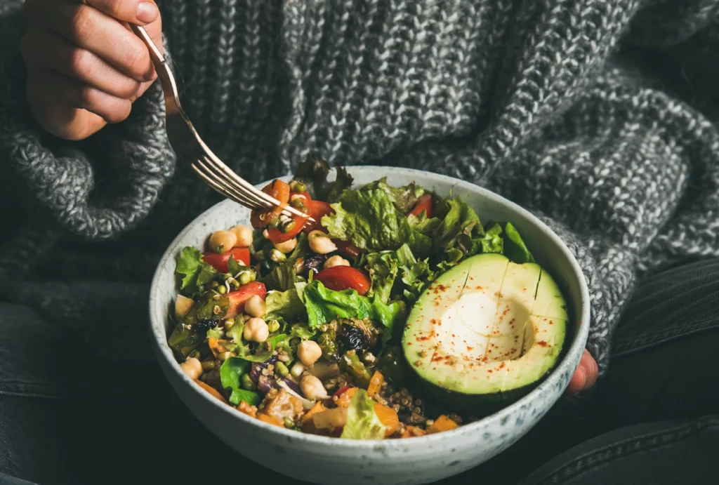 Close-up of a person in a cozy sweater eating a colorful salad bowl with greens, grains, and avocado.