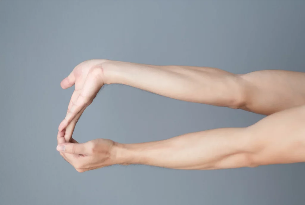 Two arms extended outward with fingers interlaced, demonstrating a hand and wrist stretch against a gray background.