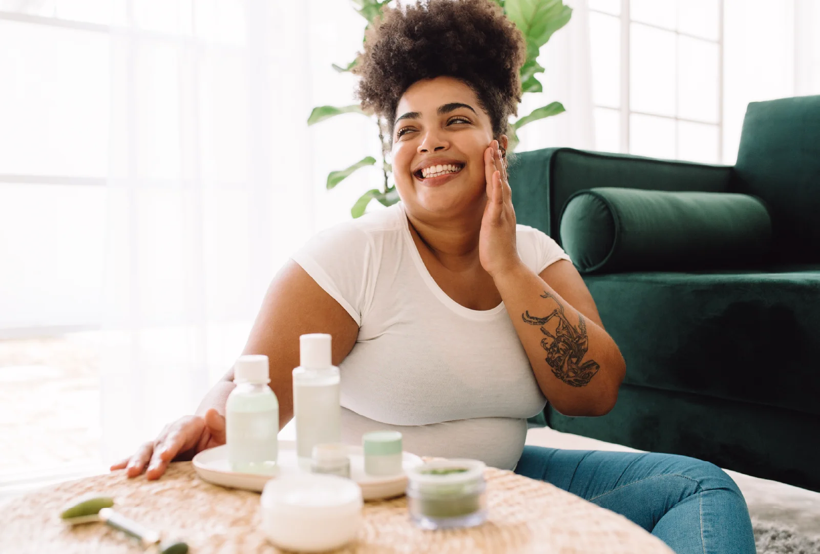 A smiling woman sitting on the floor at home, applying skincare products with several bottles arranged on a table.