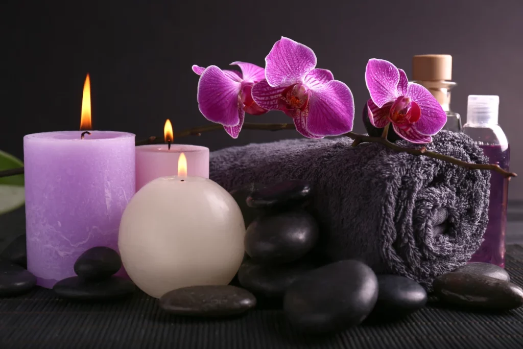 : Close up of spa items including purple candles with a black rolled-up towel, flat black rocks, and a purple orchid stem.