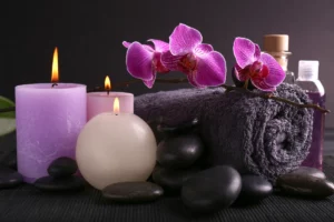 : Close up of spa items including purple candles with a black rolled-up towel, flat black rocks, and a purple orchid stem.