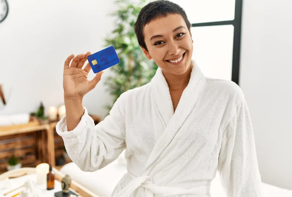 A female massage client with short hair wearing a plush white robe smiles and holds up her blue credit card in a massage room.