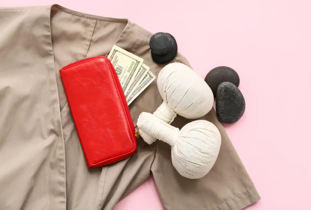 A flat-lay of a taupe massage uniform top, a red wallet with several hundred dollar bills, hot stones, and a pair of herbal massage bags on a pink background.