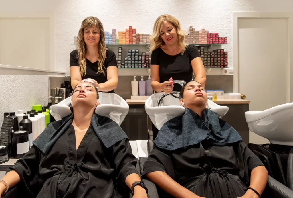 Two hair stylists smile and shampoo clients' hair at the shampoo station in a salon.
