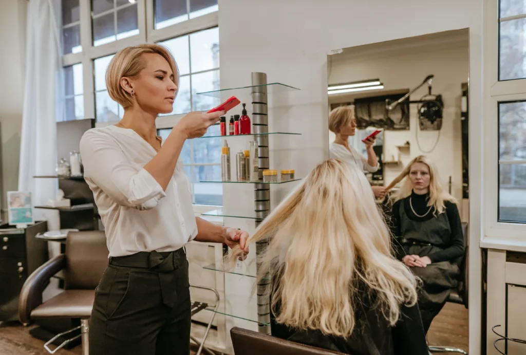A hair stylist takes a photo of a client's long blonde hair using a smartphone while the client sits in a chair in front of a vertical mirror in a salon.