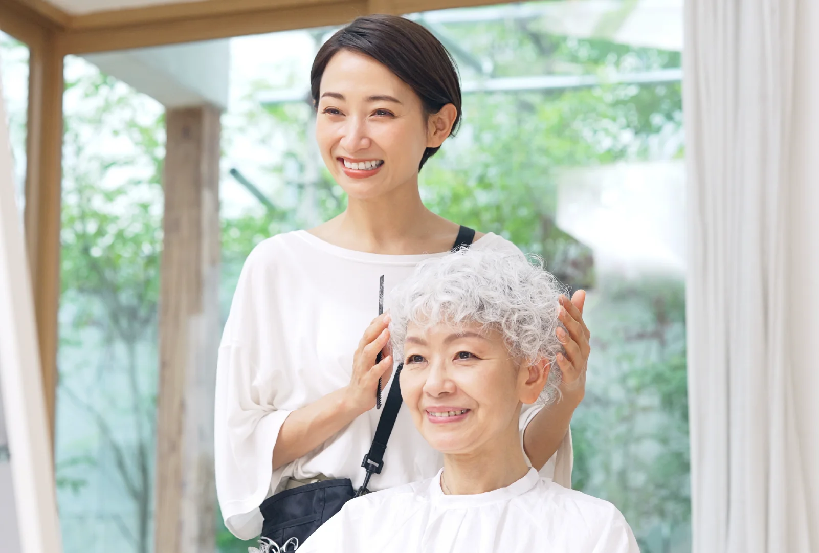 A hair stylist wearing a white blouse shapes a client's gray curly hair while the client sits in front of a mirror in a salon with a large glass window and white curtains in the background.