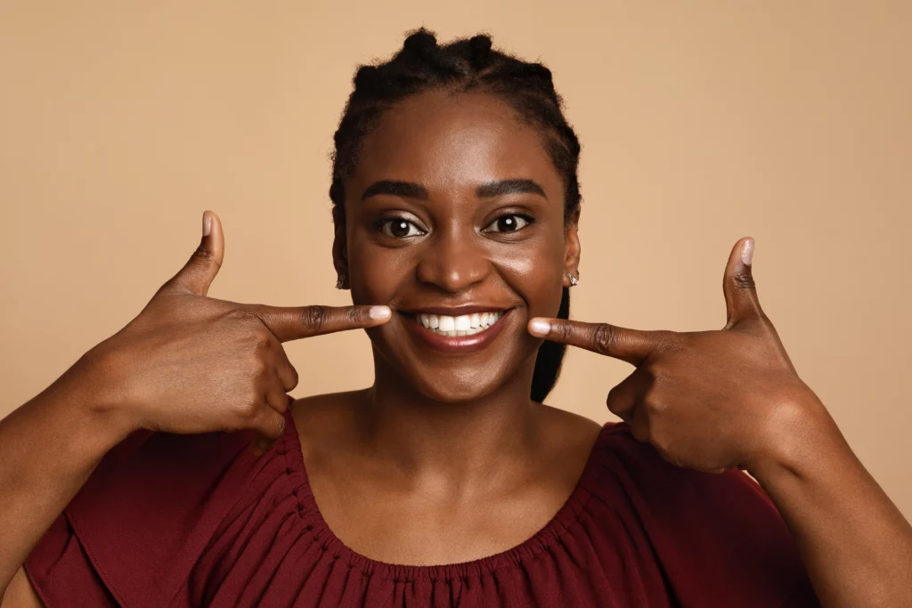 A woman wearing a burgundy top smiles and points to her teeth against a brown background.