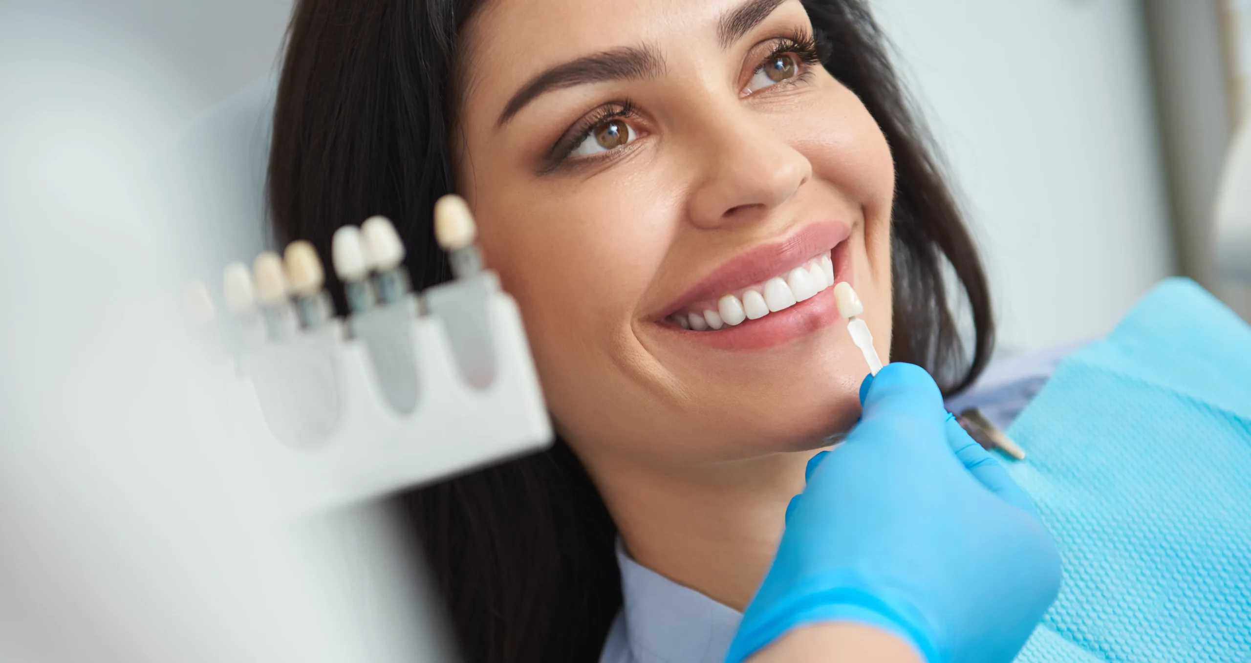 A client wearing a teal-colored paper shield smiles while a teeth whitening technician shade matches her teeth using a tooth color guide.