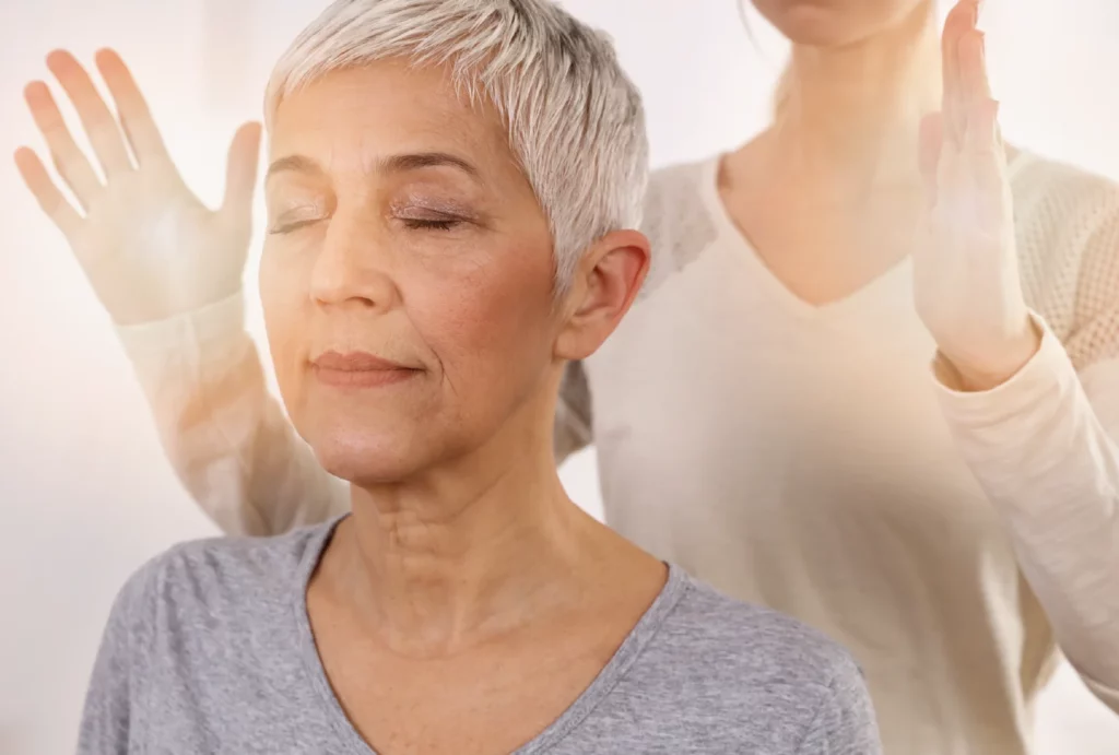 A woman closes her eyes and receives a Reiki treatment from a Reiki practitioner who holds her hands around the client's head.