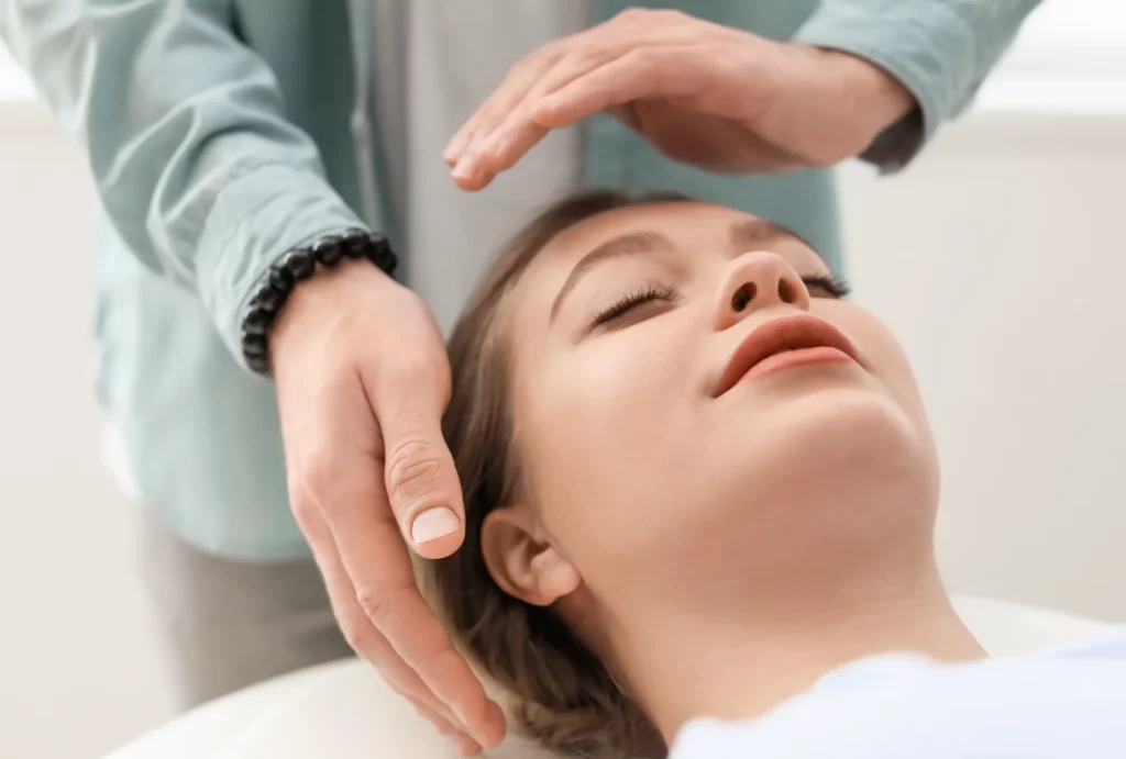 A client closes her eyes and receives a Reiki treatment from a practitioner who holds their hands slightly above the client's forehead and near their ears.