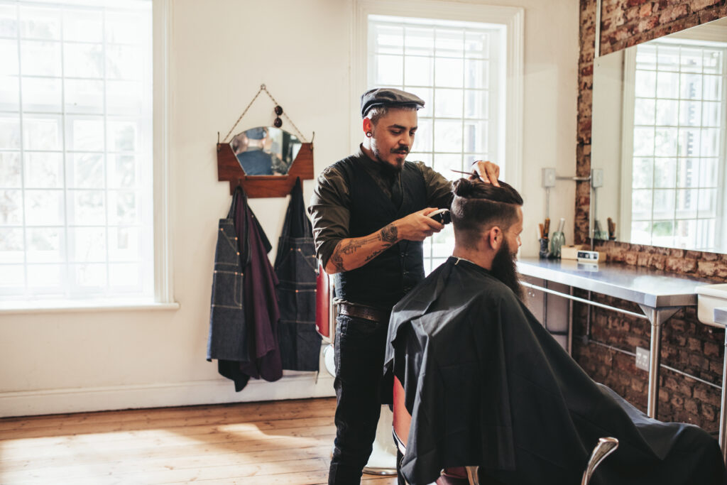 A barber with a goatee, wearing a dark vest over a dark shirt, uses a pair of clippers on a bearded client at a barber shop.