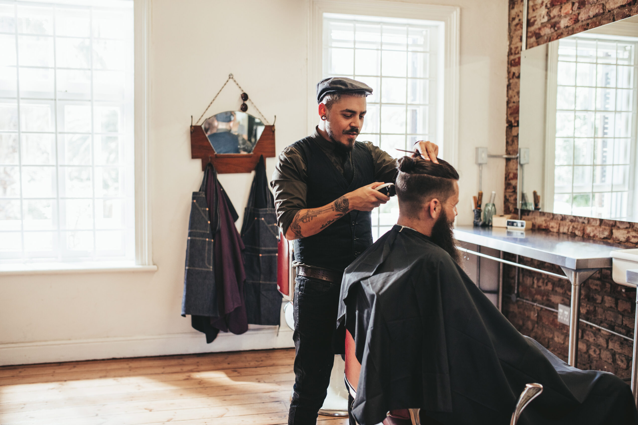 A barber with a goatee, wearing a dark vest over a dark shirt, uses a pair of clippers on a bearded client at a barber shop.