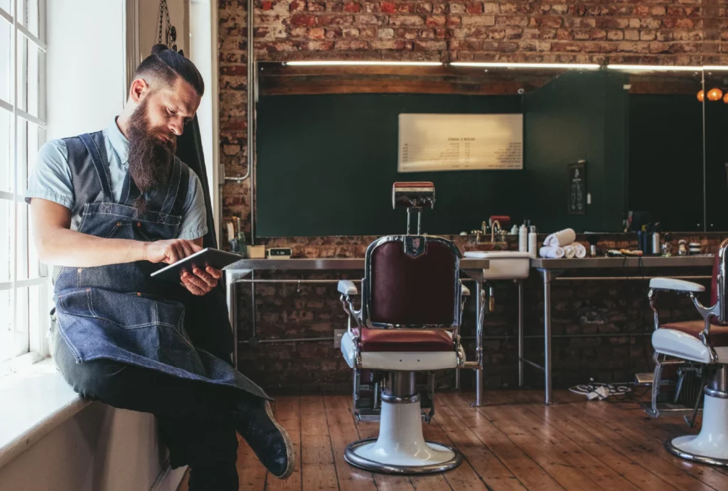 A barber works on his business tablet while sitting on a windowsill in his barbershop.