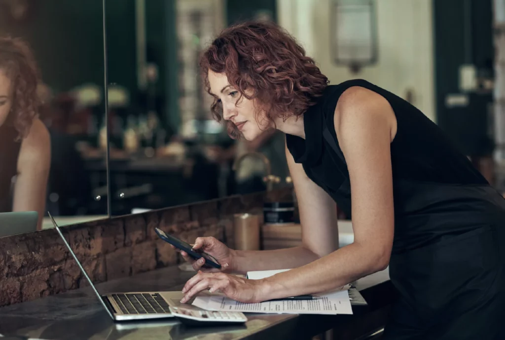 A hair stylist works on her business laptop and smartphone at a table with papers at a hair salon.