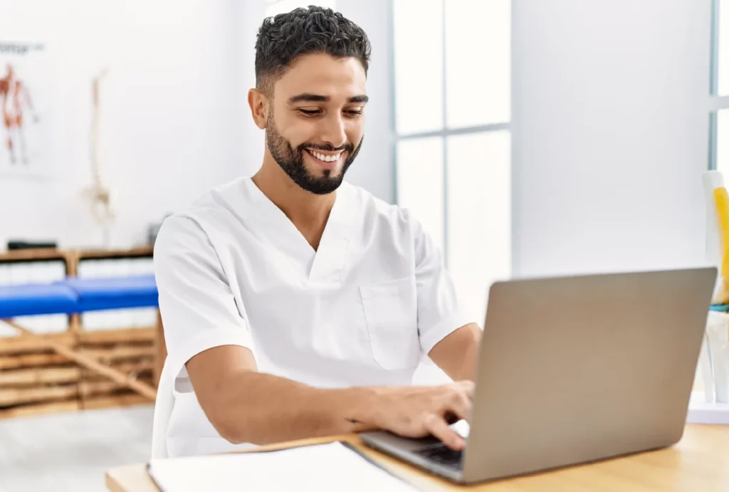 A massage therapist smiles and works on his business laptop in his practice's space.
