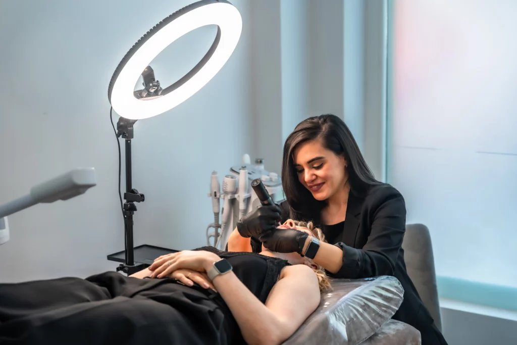 An esthetician uses a light therapy tool on a client who is lying down on a table with a large circular lamp shining from overhead.