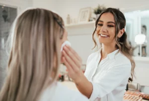 A cosmetologist wearing a white button-up shirt applies a skincare product using a cotton round to a client's cheek during a makeup application session.