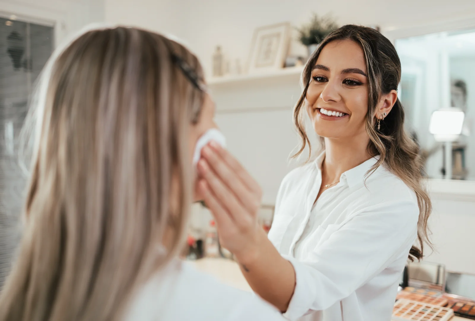 A cosmetologist wearing a white button-up shirt applies a skincare product using a cotton round to a client's cheek during a makeup application session.