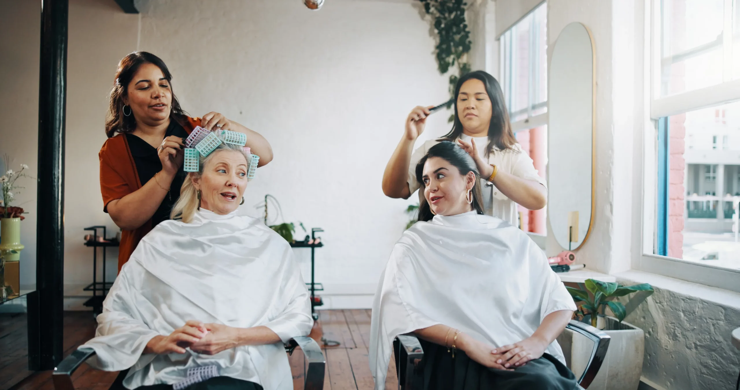 Two salon clients wearing white capes smile and chat while getting their hair done by two stylists standing behind them in a salon with white walls, open windows, and greenery.