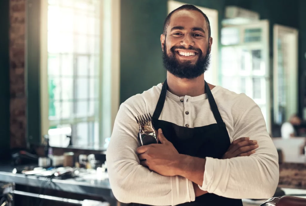 A barber smiles and holds his electric clipper in front of stations at his barbershop.
