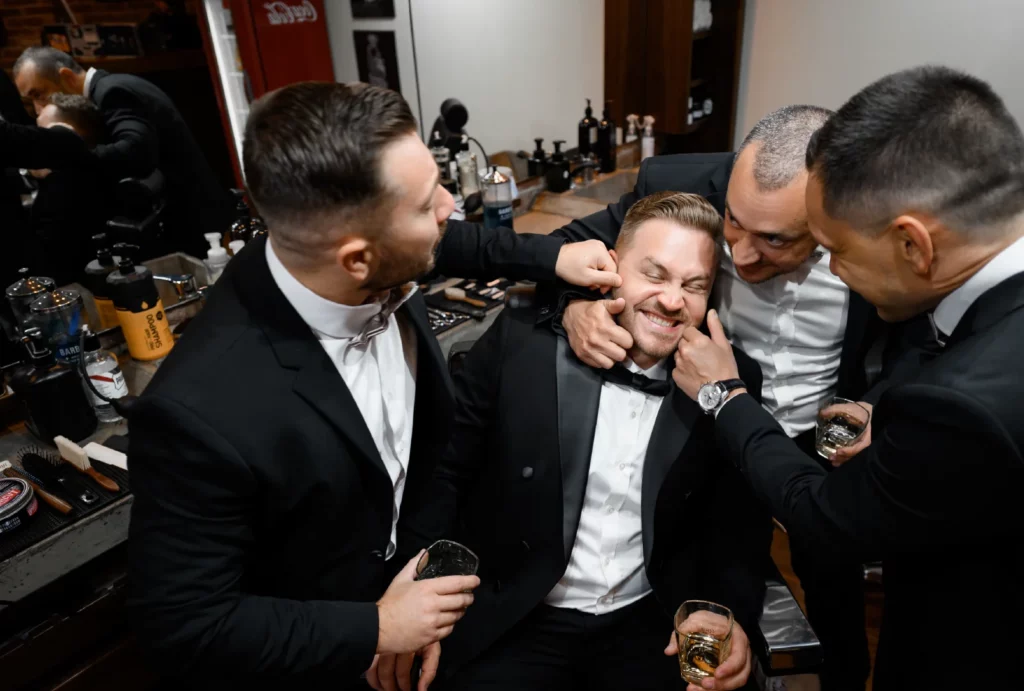 A groom's party surrounds the groom and laughs while preparing for the wedding at a barbershop.