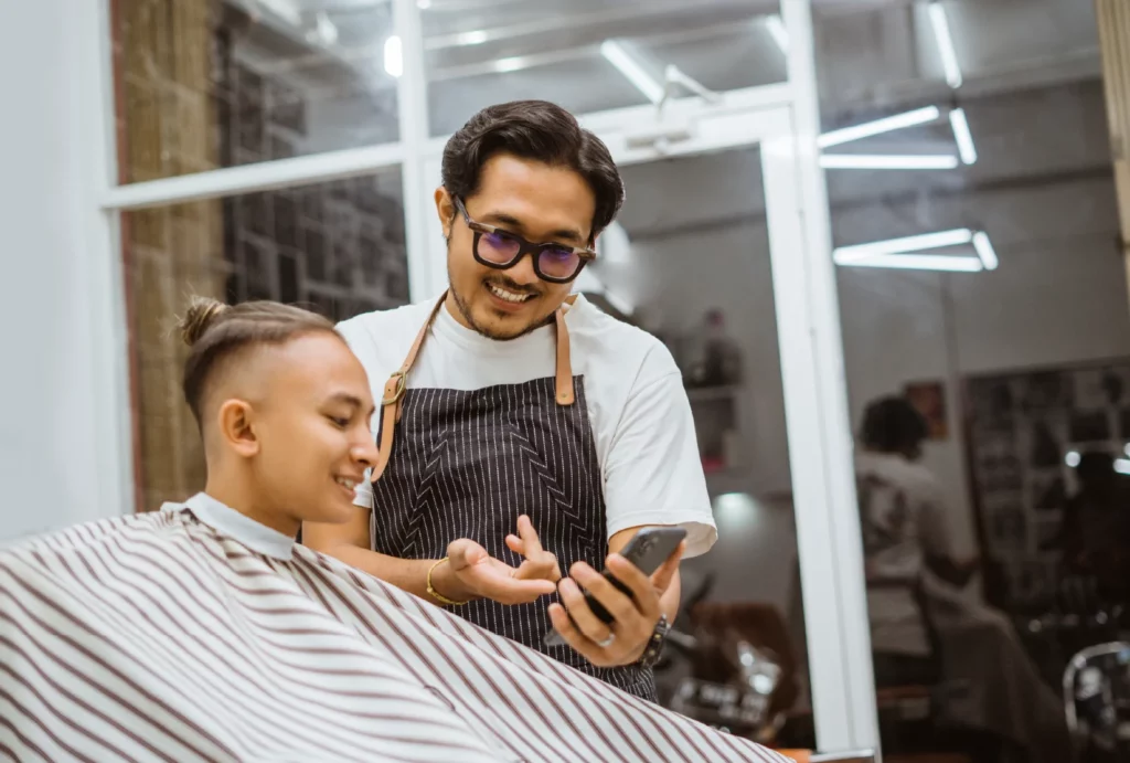A barber smiles and shows his phone screen to a client.