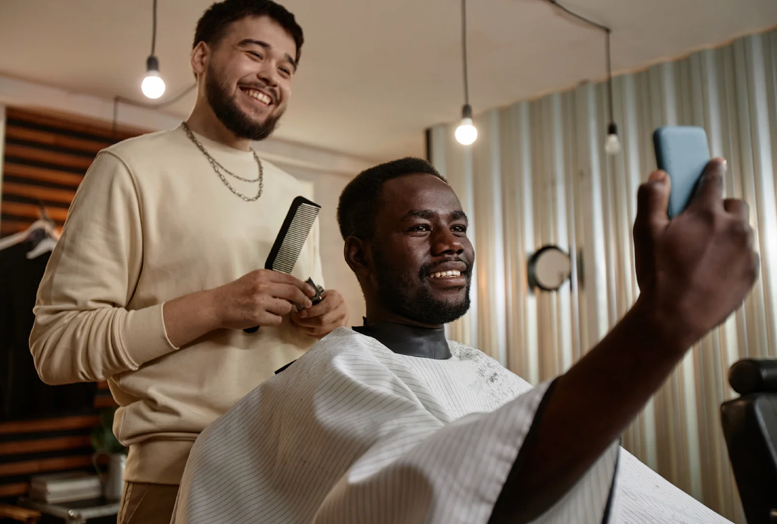 A barber smiles while standing behind a client sitting in his chair who is taking a selfie of the both of them.