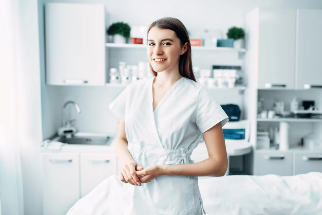 An esthetician wearing white scrubs smiles and holds her hands in front of a client bed in an esthetics space with products, shelves, and cabinets in the background.