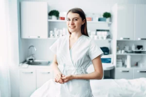 An esthetician wearing white scrubs smiles and holds her hands in front of a client bed in an esthetics space with products, shelves, and cabinets in the background.