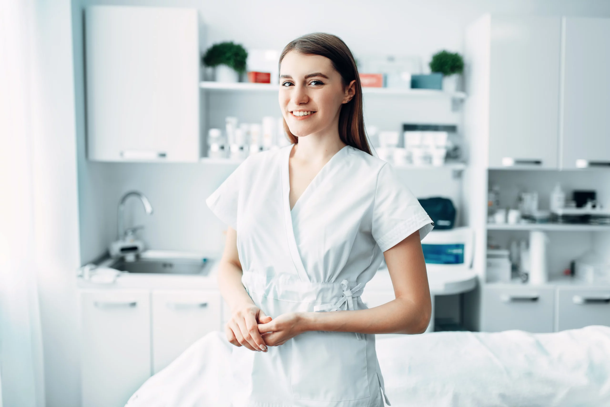 An esthetician wearing white scrubs smiles and holds her hands in front of a client bed in an esthetics space with products, shelves, and cabinets in the background.