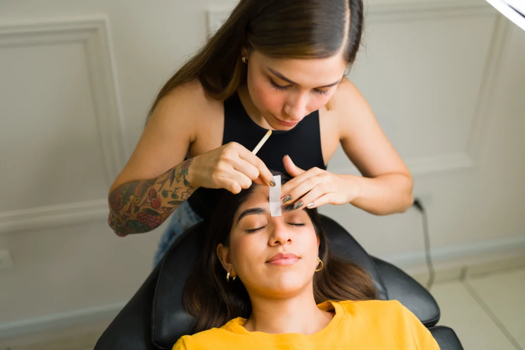 An esthetician wearing a black tank top applies eyebrow wax to a client wearing a yellow shirt who is lying down on a client bed.