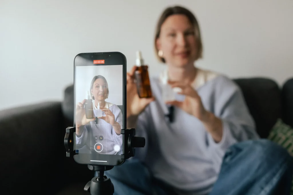 An esthetician holds up a skincare product bottle while she sits on a black sofa and films herself using a smartphone and tripod.