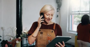 A hair stylist wearing a burgundy top and a brown apron talks on her smartphone and looks at her business tablet in a salon with potted flowers, a white wall, and a stylist in the background.
