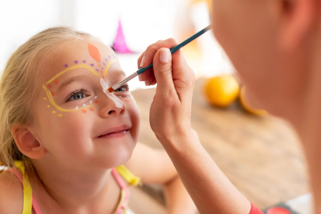 A young girl smiles while getting her face painted with yellow and white lines, and purple, orange, and yellow dots from a face painter holding a teal-colored paintbrush.