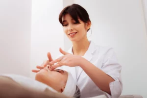 An esthetician wearing a white coat smiles and applies product to an esthetics client.
