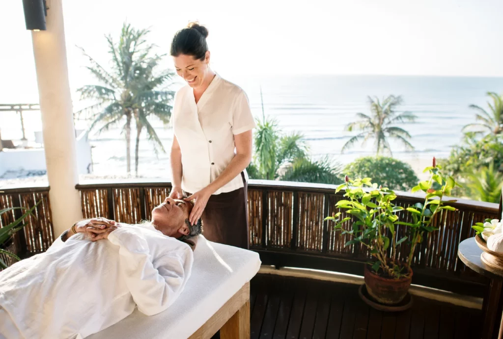 A massage therapist performing a head massage on an outdoor terrace overlooking the ocean at a spa.