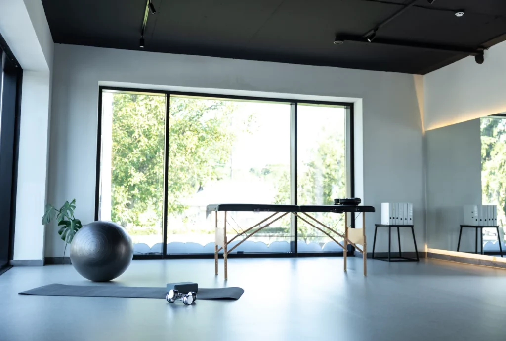 A modern physical therapy room, featuring a massage table, a physio ball, yoga mat, and a large window with light gray walls.