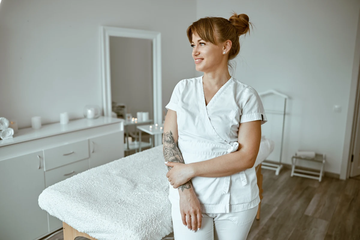 A female massage therapist with an arm tattoo and a white uniform, smiles and poses in her white massage room.