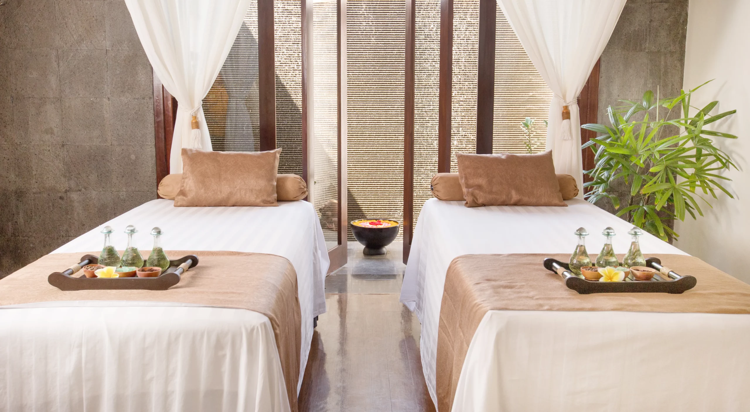 Two massage tables side by side with white and tan linens topped with trays of massage oils