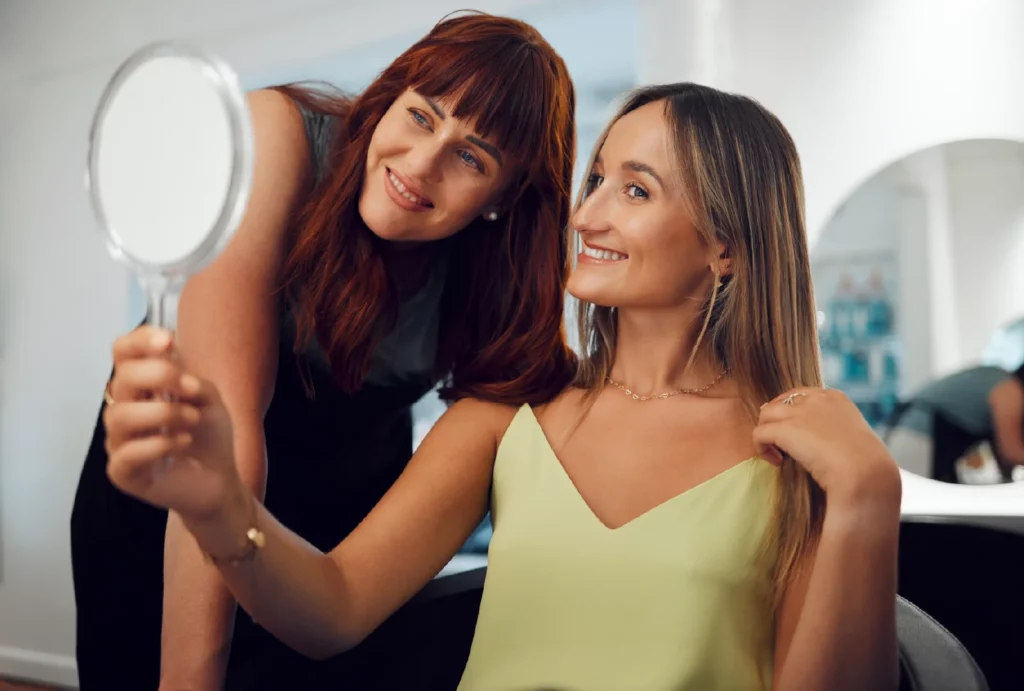 A salon client wearing a yellow-green tank admires her new hair while holding up a mirror, as the stylist leans in to see from the client's view.