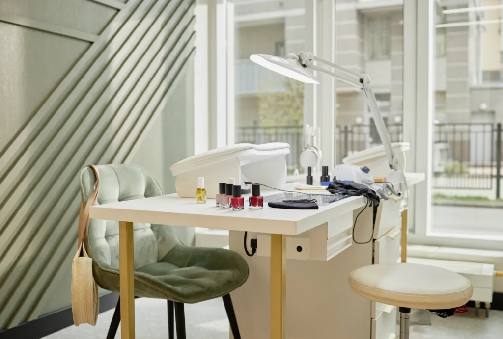 A salon manicure station with manicure supplies and a lamp on the table, a green chair with a purse hung over the side, a green accent wall, and a glass window.