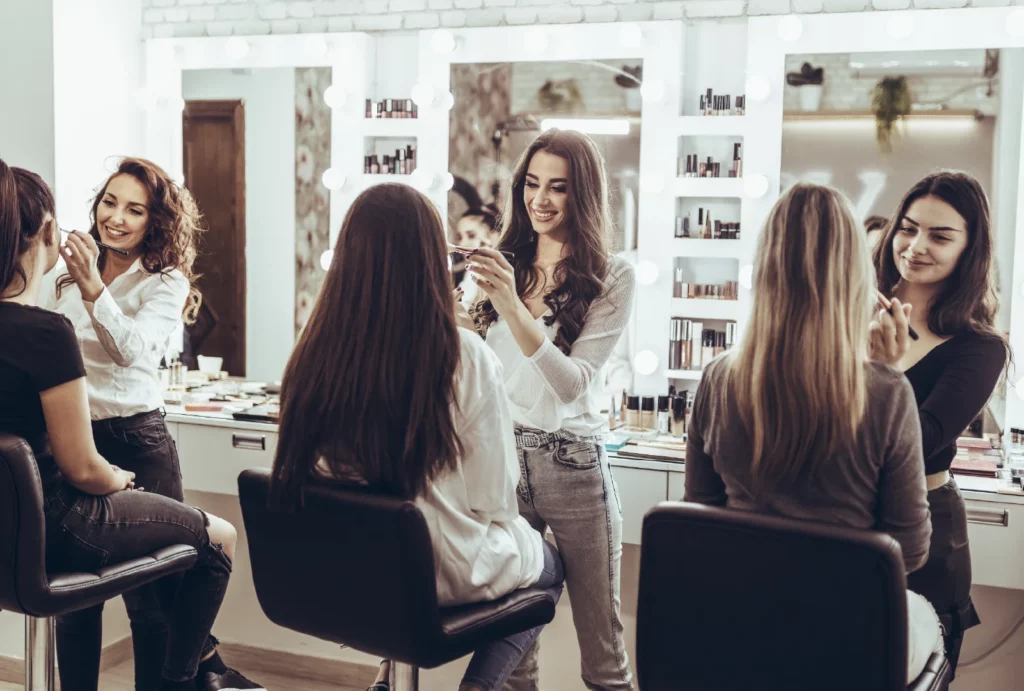 Three makeup artists apply makeup to three clients in a salon with brightly lit mirrors, makeup products on the stations, and black salon chairs.