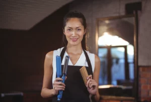 A hair stylist wearing a white sleeveless top and a black apron holds a blue hair straightening tool and a brown comb while smiling in a salon with a red-brick wall and a mirror in the background.