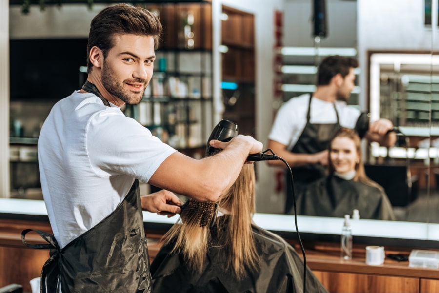 man looking back at camera as he blow dries clients hair in salon