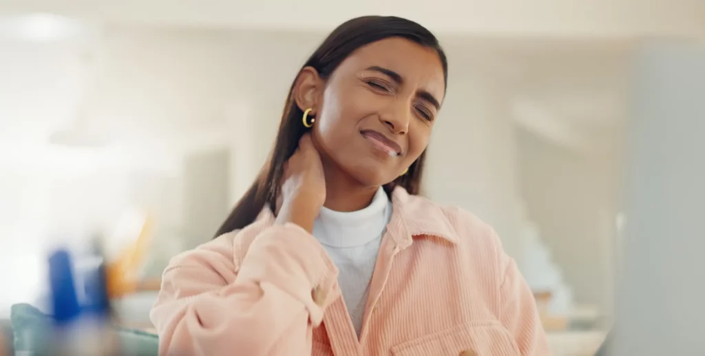 A woman with straight brown hair, wearing a white mock neck top and a pink jacket, grimaces and holds her neck in pain.